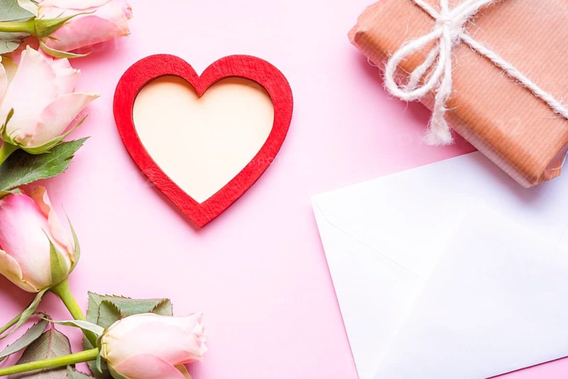 Valentine day theme image with a red heart with copy space in the middle, surrounded by pink roses, an envelope and a gift wrapped in brown paper, on a pink background