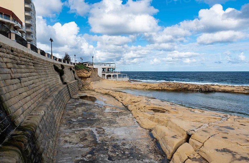 Saint Julian, Malta, 01 07 2022: Low angle view over the water, rocks and sand at the beach with the harbor in the background