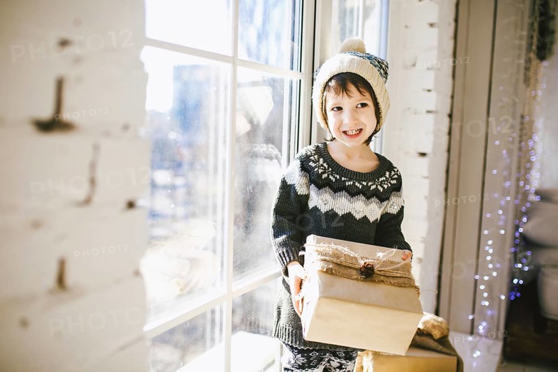 Child sitting at the window on a sunny Christmas day, making presents wrapped in boxes in paper. dressed in knitted warm woollen clothes and hat. Inside the house with a stylish interior
