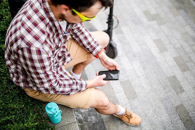 Caucasian man uses modern technologies Internet and cellular communication on smartphone while sitting on bench in city near electric scooter. Ecology and environmentally friendly urban transport