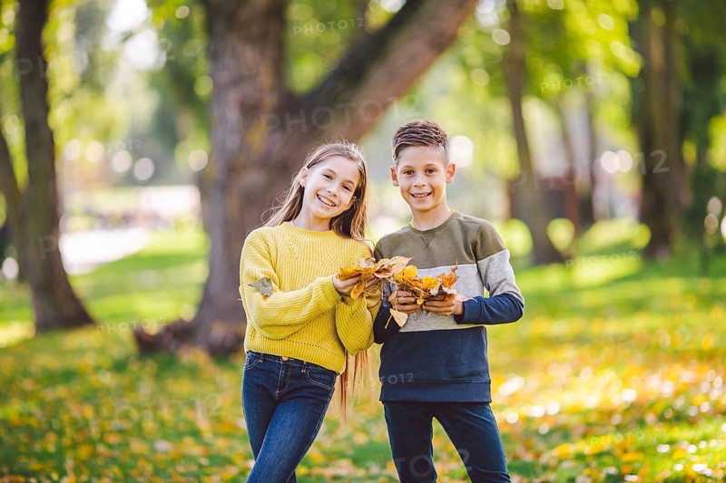 Happy Caucasian siblings enjoying in autumn day in park, sunny weather. Kids having fun holding fallen yellow leaves in nature. Funny twins in autumn park. Autumnal mood. Teenagers posing embracing