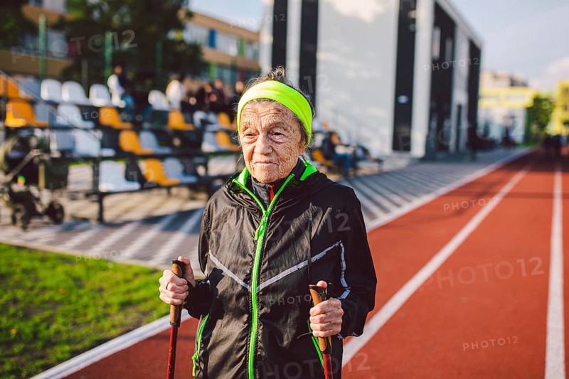 Active rest of the elderly theme. Sports and health in retirement. Caucasian very old woman with deep wrinkles doing Nordic walking exercises with sticks outside in the city stadium