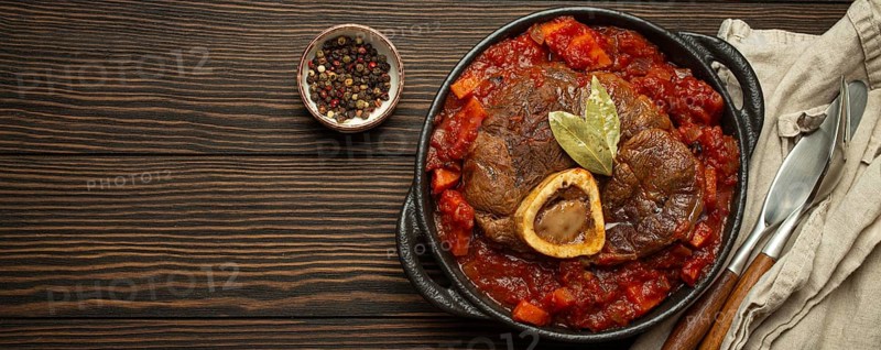 Traditional Italian dish Ossobuco all Milanese made with cut veal shank meat with vegetable tomato sauce served in black casserole pan top view on rustic brown wooden background, copy space, food photography, food photography