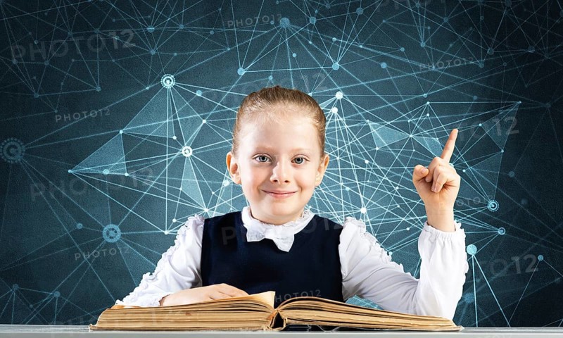 Smiling little girl sitting at desk with open book. Research and education in school. Happy schoolgirl in school wear finger pointing up. Clever girl reading book on deep blue plexus background