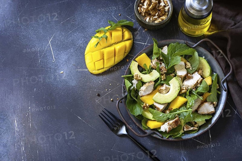 Healthy diet salad with arugula, mango, avocado, chicken grill, walnuts and vinaigrette dressing on a dark stone countertop. Copy space