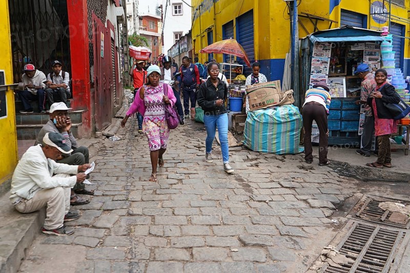 Antananarivo, Madagascar - April 24, 2019: Two unknown young Malagasy girls walking cobblestone road, more people around at their stalls, or sitting on ground - typical scene in streets of Tana city, Antananarivo, Madagascar
