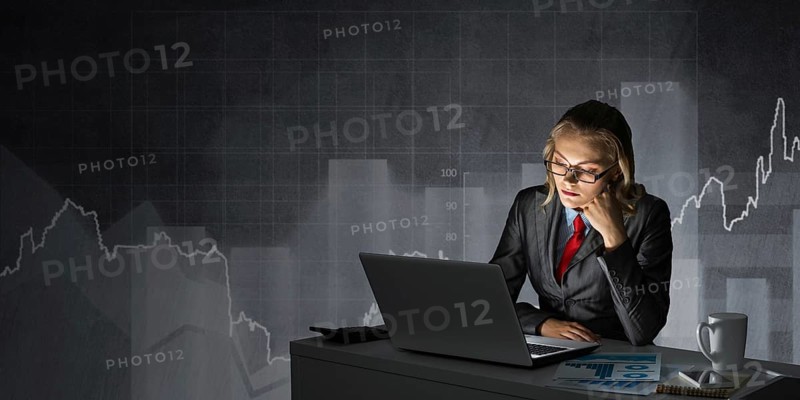 Attractive young businesswoman in glasses sitting at office desk and working at laptop. Female accountant in business suit and tie busy at work in evening. Financial analysis and planning