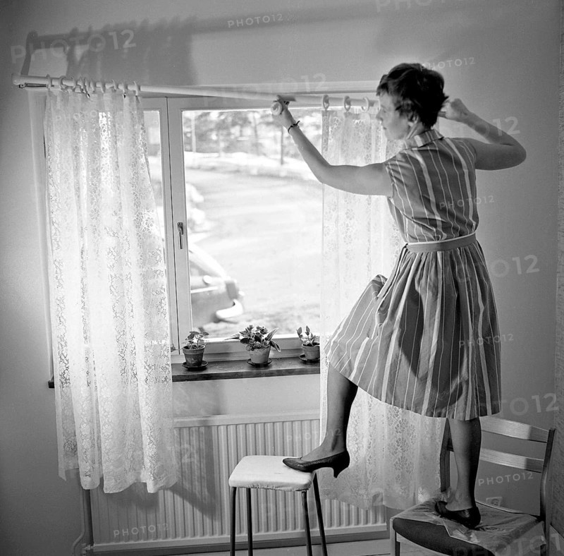 1960s home decorating. A woman at home putting up the curtains in the room window. All by herself she balances on a chair and a stool to manage it. Sweden 1966