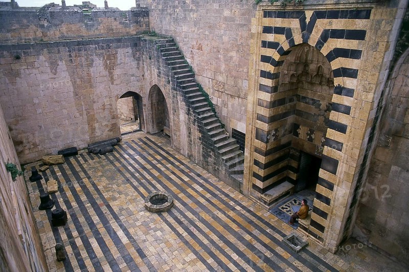 The Citadel of Aleppo is a large medieval fortified palace in the centre of the old city of Aleppo, northern Syria. It is considered to be one of the oldest and largest castles in the world. Usage of the Citadel hill dates back at least to the middle of the 3rd millennium BCE. Subsequently occupied by many civilizations including the Greeks, Byzantines, Ayyubids and Mamelukes, the majority of the construction as it stands today is thought to originate from the Ayyubid period.<br/><br/> Aleppo, the second city of Syria and quite possibly the longest continually inhabited settlement in the world, is of venerable age. So old, indeed, that its Arabic name, Halab, is first mentioned in Semitic texts of the third millennium BCE. Situated in the north-west of the country, just a few kilometres from the Turkish frontier, Aleppo is located at the confluence of several great trade routes and, as a city of commerce, has always been rich