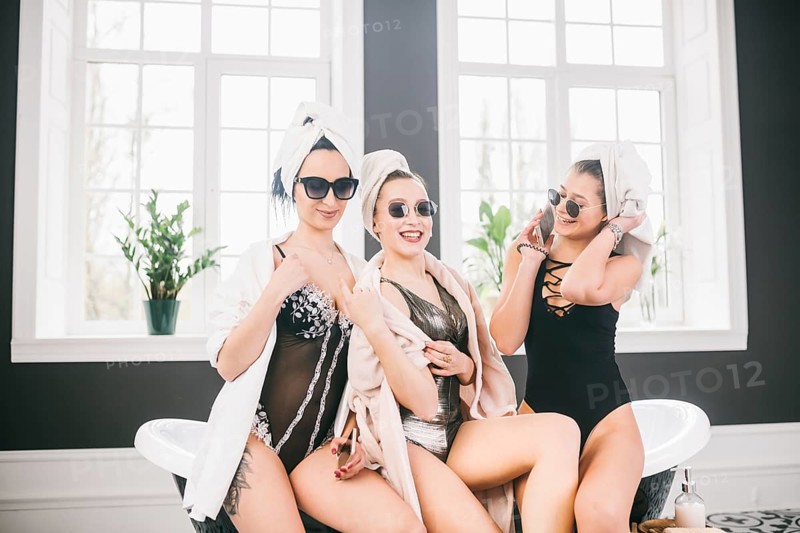 Theme female friendship. Party girlfriends, relaxation and spa procedures, hen party. Three Caucasian beautiful women sitting inside on bathroom in sunglasses, head towel and bathrobe, underwear