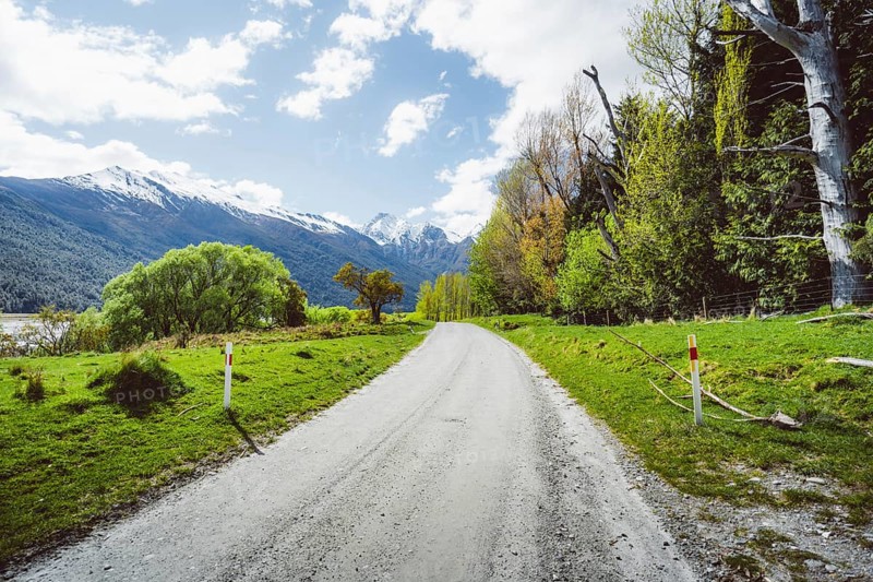 A rural road leads through a green landscape with snow-capped mountains in the background, Cattle Flat, Wanaka, New Zealand