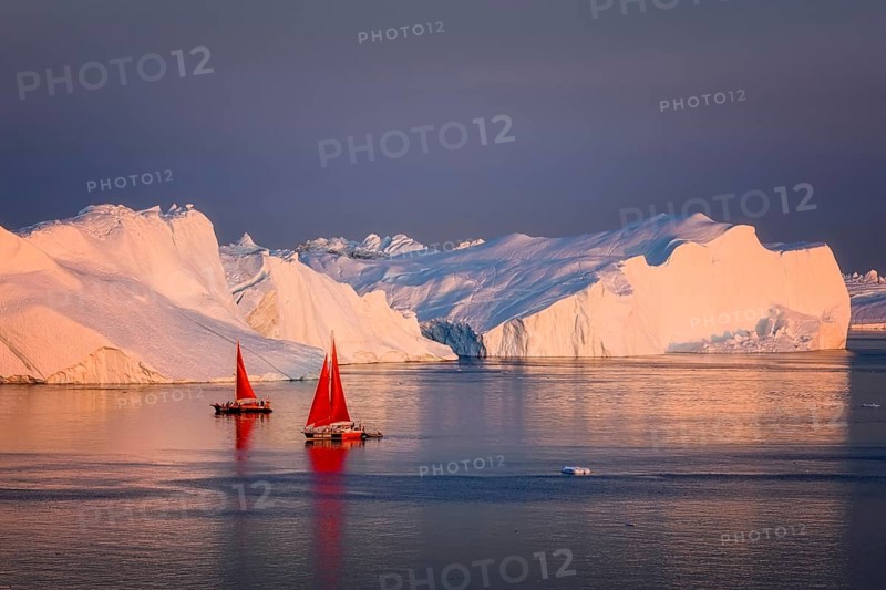 Large glaciers in the bay by the rocky shore in the light of the setting sun with fog over the water with red boat