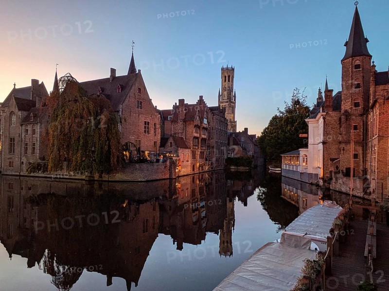 Charming canal at sunset with medieval buildings and a distant tower reflected in the calm water. Bruges, Belgium