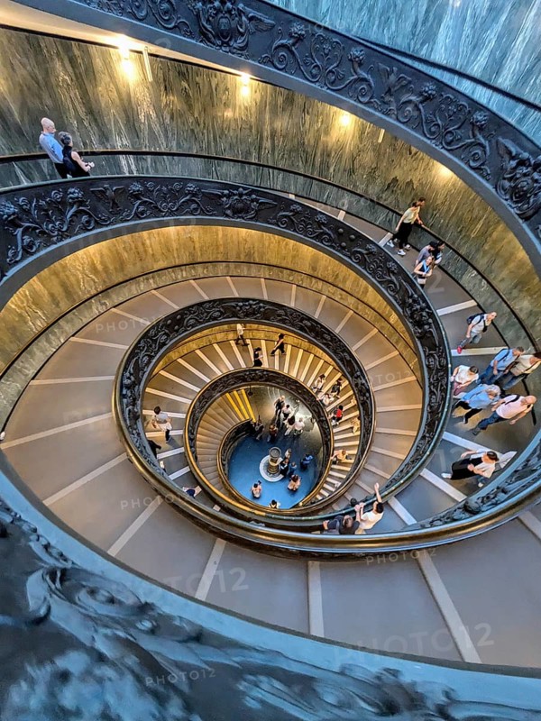 Aerial view of a busy spiral staircase with people ascending and descending, highlighting the ornate railings. The Vatican