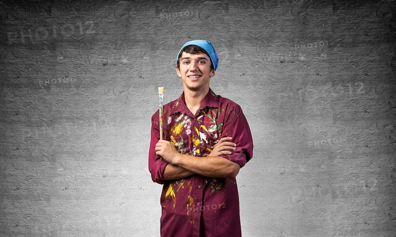 Young happy painter artist holding paintbrush. Portrait of painter in dirty shirt and bandana standing on gray wall background. Creative hobby and artistic occupation. Art school courses concept