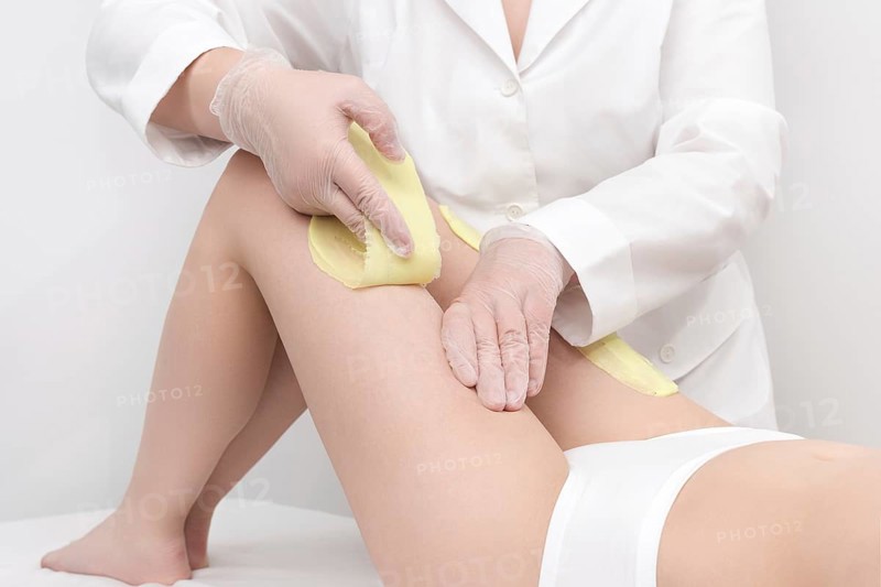 Unrecognizable beautician in white coat removal hair on women legs. Depilation procedure with green hot wax in professional beauty salon while woman lying down on couch. Part of photo series