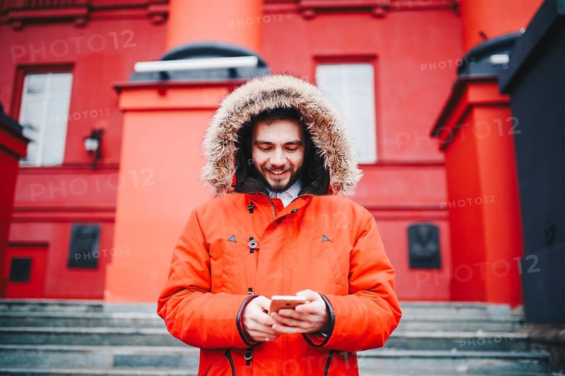 Handsome young male student with toothy smile and beard stands on red wall background, facade of educational institution in red winter jacket with hood with fur, Uses finger on screen of mobile phone