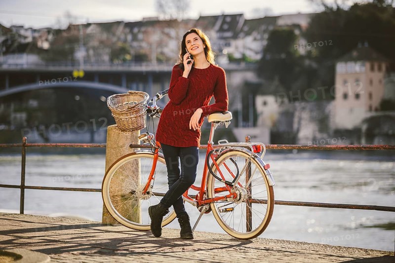 A young female traveler enjoying stands next to a bicycle near a river in the city of Basel. Traveling in Switzerland. Young caucasian woman talking by cell phone on bridge in european city