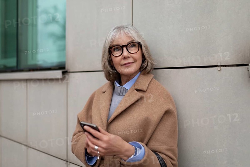 Eine reife Frau in einem hellbraunen Mantel steht an einem modernen Gebäude und überprüft ihr Telefon, während sie einen Moment der städtischen Ruhe genießt