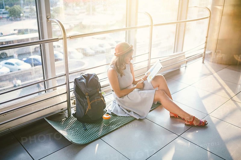 Waiting, delayed transport in the terminal of the airport or train station. Young caucasian woman in dress and hat sits on tourist rug with backpack near window to room and looks into tourist map