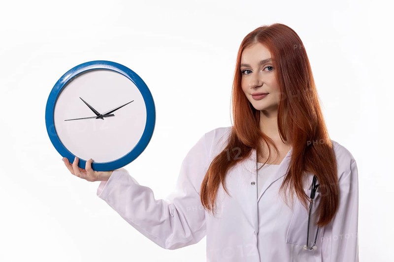 A female healthcare worker in a lab coat holding a clock, symbolizing time management in healthcare