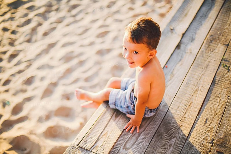 Little Caucasian boy child sitting on wooden pier sandy beach, summer time, sea holiday near water. The theme is the flow of time, a short life, the meaning and purpose of existence. Meaning of life