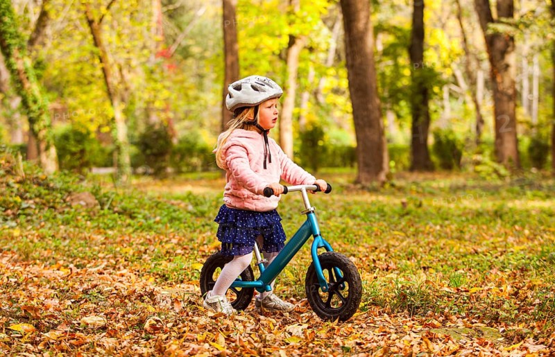 Side view preschool girl in protective helmet riding blue balance bike at the autumn park. Lovely girl in skirt on bike among fallen leaves
