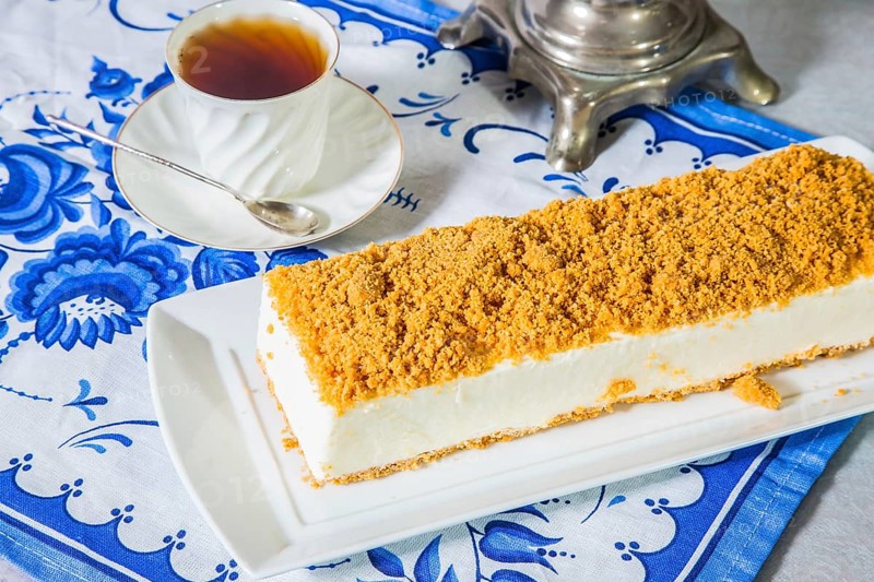 Professional bakery. Exquisite white cheesecake, sprinkled with sweet orange crumbs. The background is samovar and porcelain white cup with hot tea on blue kitchen towel