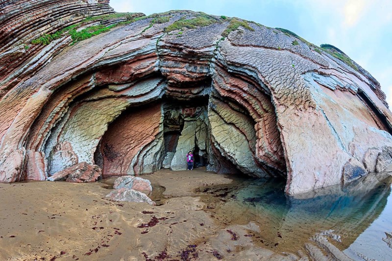 Picturesque huge cave on the beach. Low tide in the Atlantic. Flysch - unique landscape of underwater rocks exposed at low tide. Woman in pink jacket photographs Playa Zumaya