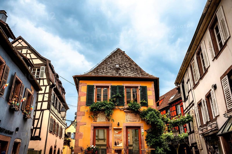 Characteristic half-timbered house with colourful shutters and floral decorations under a cloudy sky. Alsace France