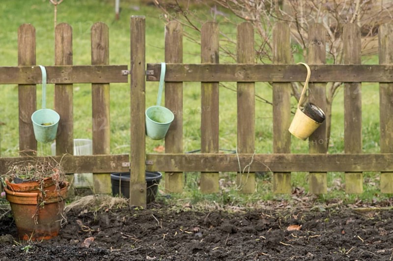Three empty buckets hanging on a weathered wooden fence in a garden, with a terracotta pot and bare soil in the foreground