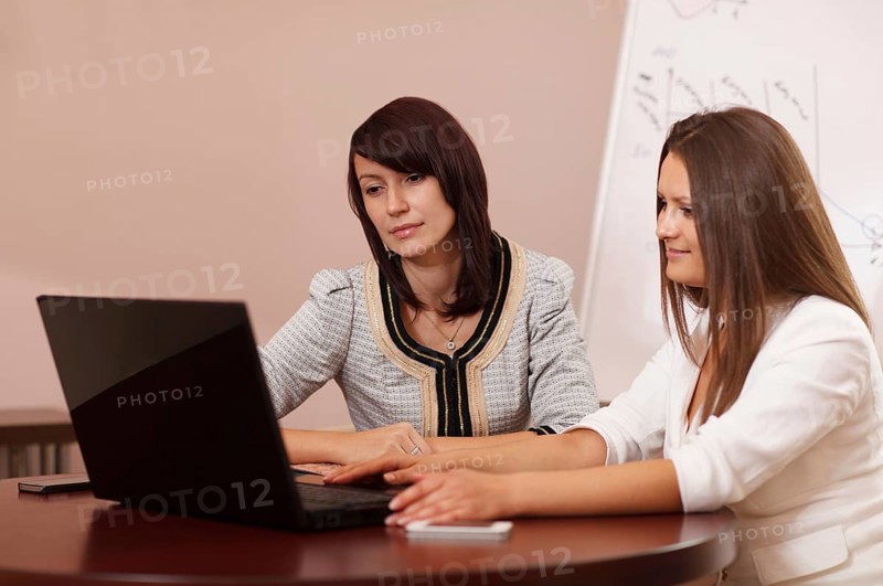 Two attractive stylish young women sitting at a table with a laptop computer reading something on the screen together while browsing the internet