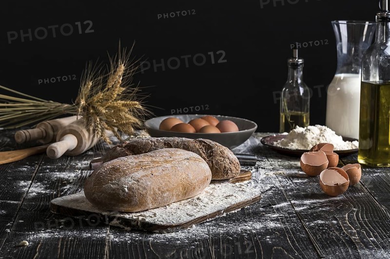 Beautiful still life with different kinds of bread, grain, flour on weight, ears of wheat, pitcher of milk and eggs on black background