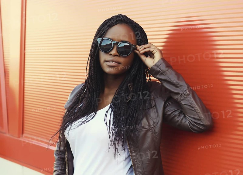 Fashionable portrait of young African woman in casual clothing, stylish modern black girl in glasses with dreadlocks posing in the city on red background