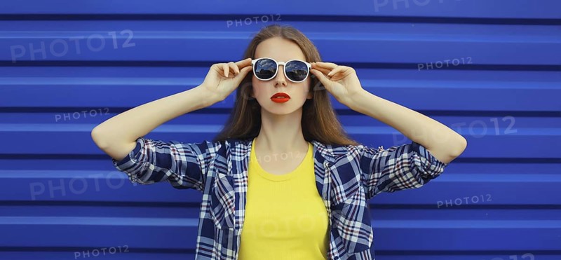 Portrait of modern stylish young woman in white glasses, casual clothing posing on blue wall background