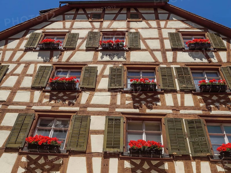 Detailed view of a half-timbered façade with flower boxes full of red flowers and shutters against a blue sky, Meersburg, Lake Constance, Germany