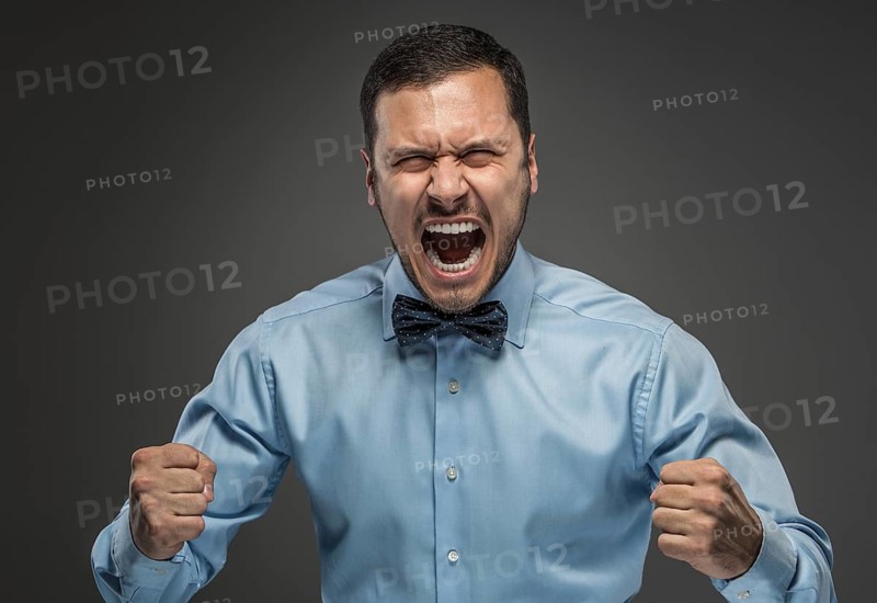 Portrait of angry upset young man in blue shirt and butterfly tie with fists up yelling isolated on gray studio background. Negative human emotion, facial expression. Closeup