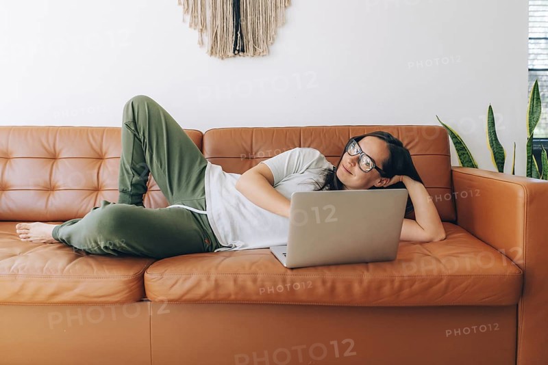 Junge glückliche Frau mit Brille, die zu Hause auf der Couch liegt und an ihrem Laptop arbeitet