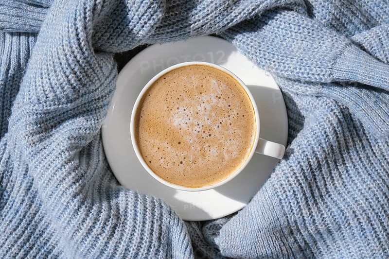 White cup of morning coffee on blue sweater background. Cozy home autumn concept. Aesthetics blog lifestyle. Still life concept. Cappuccino or latte hot drink. Top view flat lay