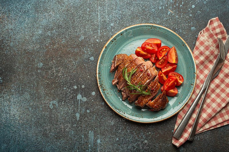 Delicious roasted sliced duck breast fillet with golden crispy skin, with pepper and rosemary, top view ceramic blue plate served with cherry tomatoes salad, concrete rustic background, copy space, food photography, food photography