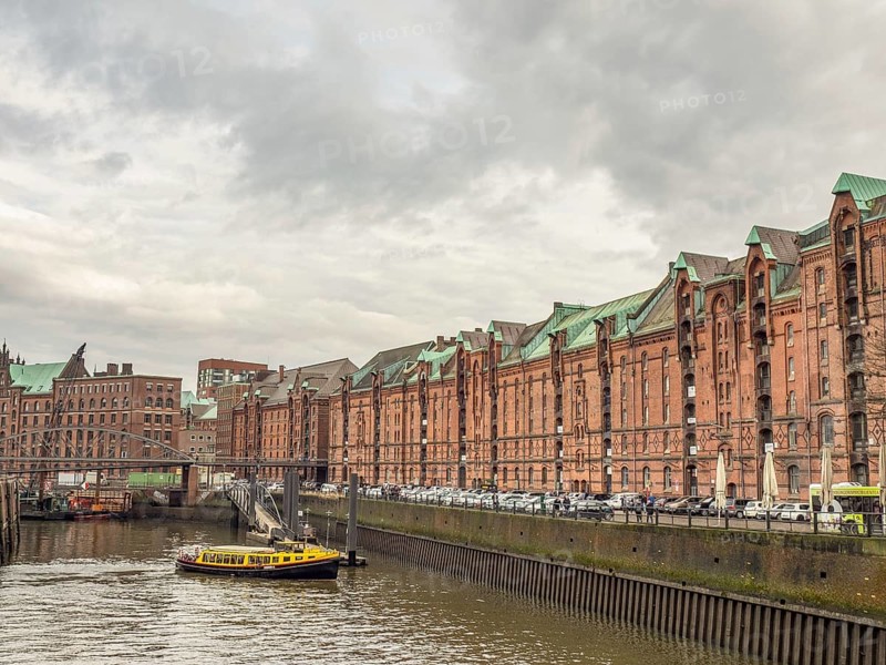 Yellow boat on a river next to historic brick-red buildings and a metal bridge under a cloudy sky, Hamburg, Germany