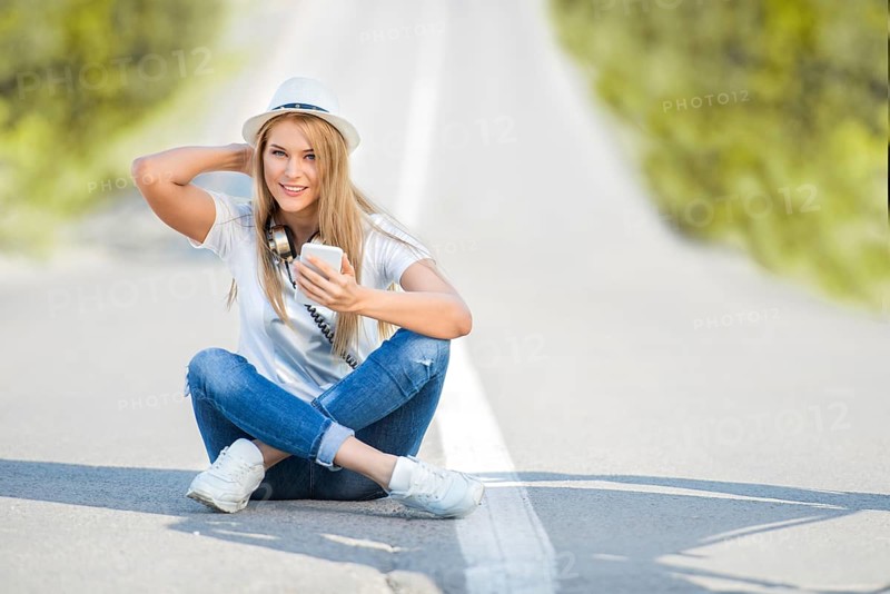 Happy young woman with vintage headphones around her neck, surfing the internet on her smartphone and sitting on a divider against the backdrop of a street