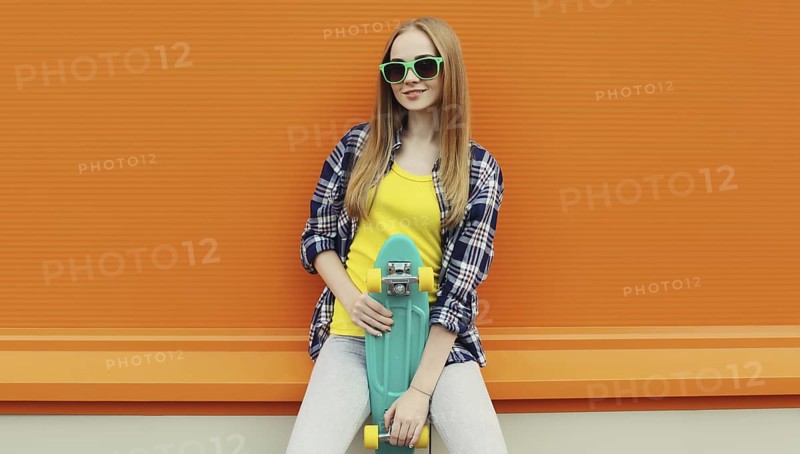Summer portrait of happy cheerful stylish young woman with skateboard, teenager girl in colorful clothes, glasses posing on orange background on city street
