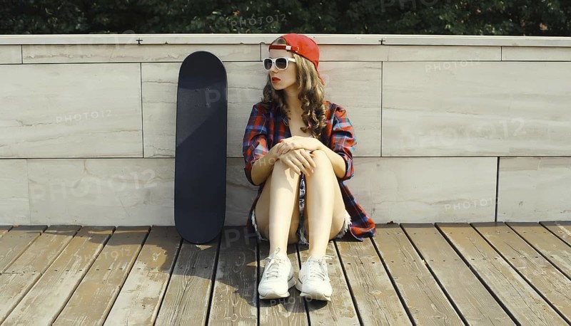 Stylish young woman sitting with skateboard in casual clothes looking away, modern teenage girl posing in the city