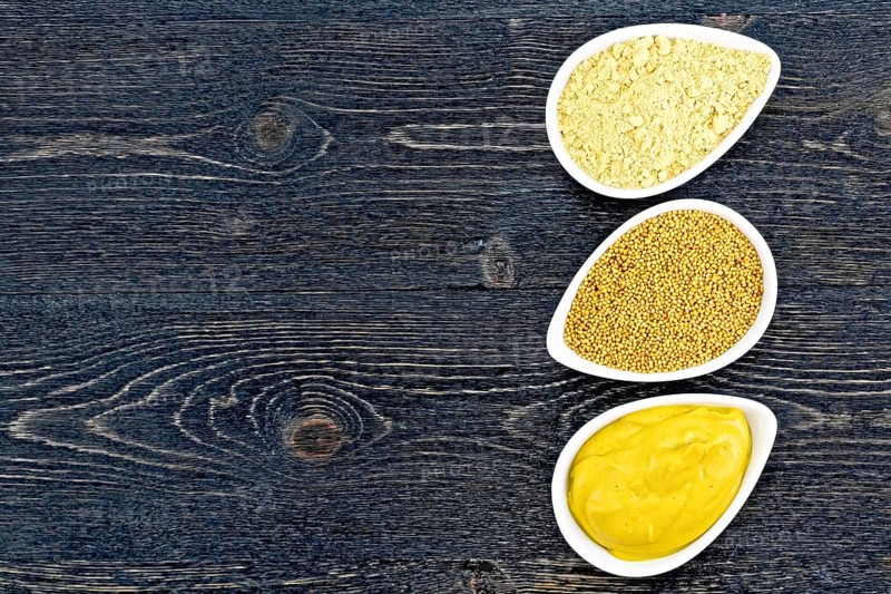 Mustard sauce, seeds and mustard powder in three saucepans on the background of a black wooden board from above