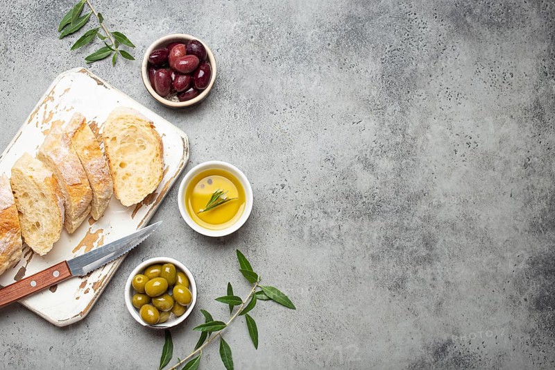 Sliced fresh ciabatta on cutting kitchen board, green and brown olives, olive oil with rosemary, olive tree branches on gray concrete stone rustic background from above copy space, food photography, food photography