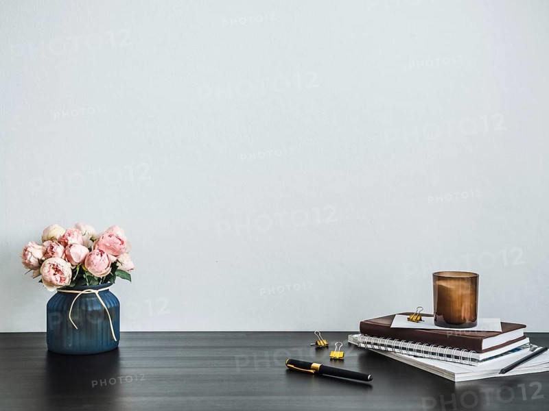 Home office desk with copy space. Pink flowers, scented candle, stack of books on black tabetop. Copy space for text. Home office desk workspace. Empty white room with wooden table