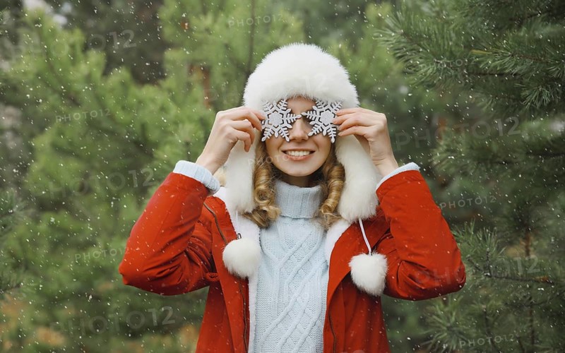 Winter day, happy smiling young woman with snowflakes decoration toys having fun enjoys snow, warm weather against Christmas tree, joyful girl in white hat standing in snowy forest
