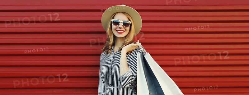 Stylish beautiful happy smiling young woman posing with shopping bags in summer straw hat on red wall background