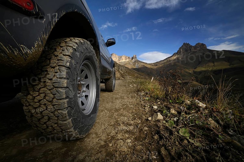 Close-up A large off-road all-terrain car wheel stands on a rough road in the mountains against the backdrop of rocks. Off-road tours and off-road travel concept